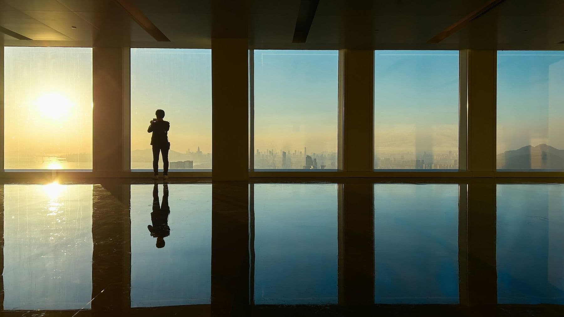 Lone figure silhouetted against floor-to-ceiling windows overlooking a city at sunrise
