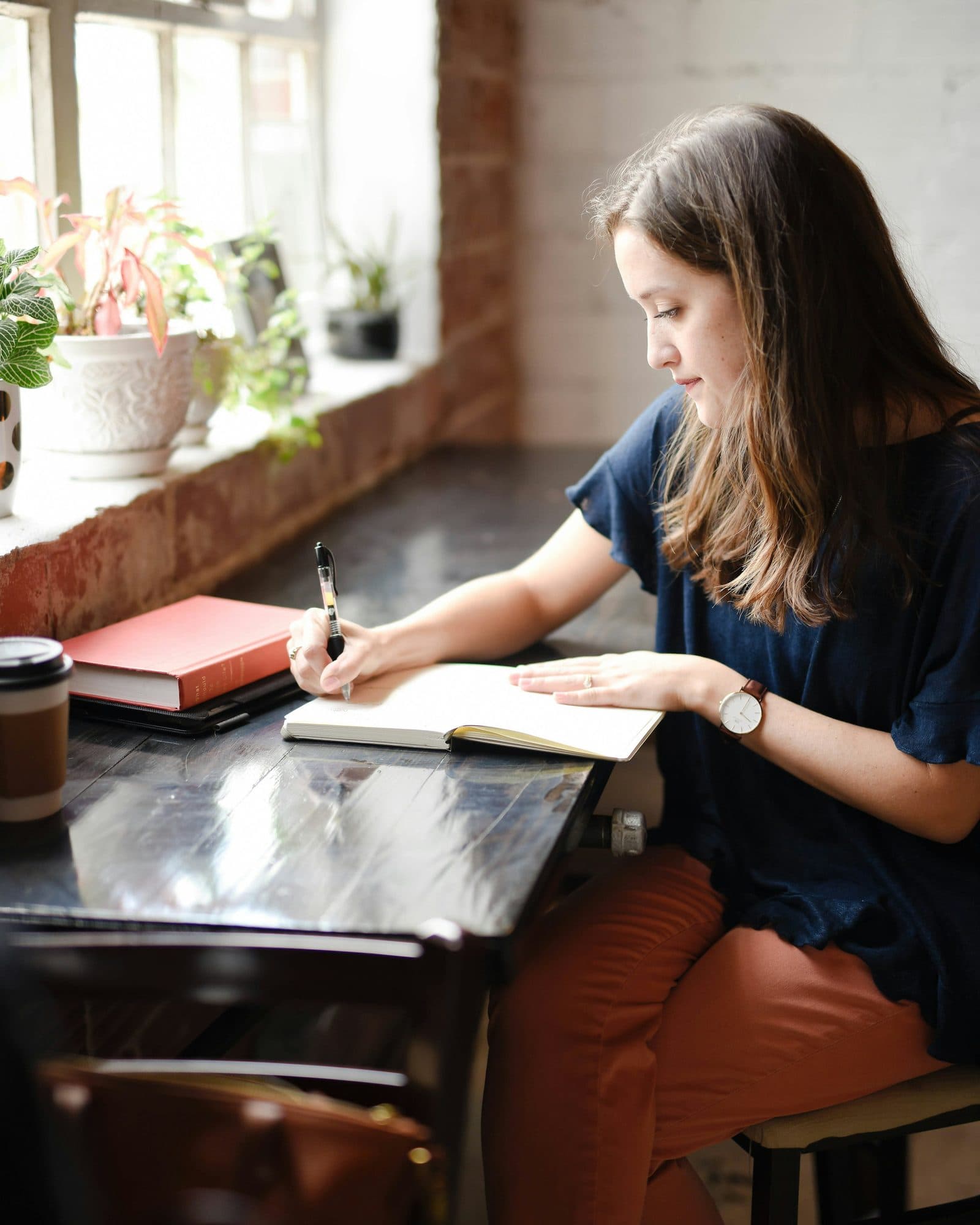 Woman annotating a notebook at a window-lit desk with coffee and books