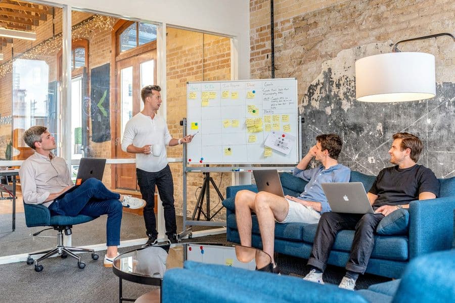 Sales team huddle with a presenter at a whiteboard of sticky notes in a loft office