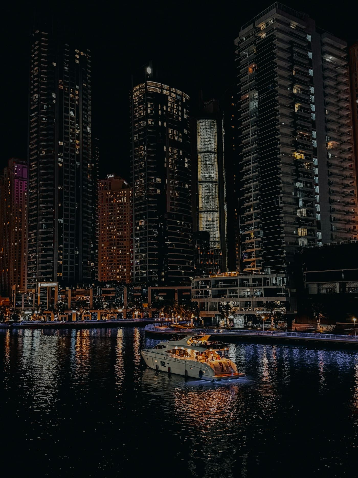 A yacht cruising through Dubai Marina at night between lit towers